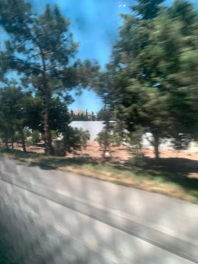 The image captures a blurred scene, likely taken from a moving vehicle, showing trees against a bright blue sky. In the background, a white wall is visible, with some foliage and a building with a curved roof peeking over it. The foreground is dominated by a blurred grey surface, possibly a road or a window with reflections. The trees are evergreen with dark green foliage and brown trunks. The overall impression is that of a sunny day, possibly in a suburban or rural area, given the presence of trees and the absence of dense urban structures. The location is specified as Mardakan, Azerbaijan.