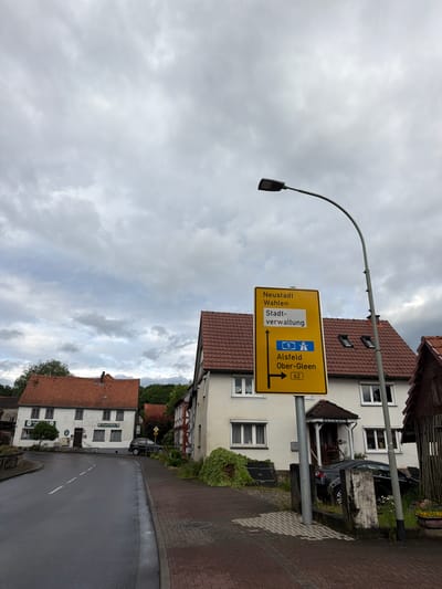 The image shows a street scene in Kirtorf, Germany. A yellow directional sign is prominently displayed, indicating directions to Neustadt, Wahlen, Stadtverwaltung, Alsfeld, and Ober-Gleen, with the latter marked with a route number 62. A street lamp with a curved pole is situated to the right of the sign. In the background, there are traditional German houses with red tile roofs and white walls. One building on the left has a sign that reads "Gaststätte," indicating it is a restaurant or guesthouse. A black car is parked on the right side of the road. The sky is overcast with heavy clouds. The road is wet, suggesting recent rain. The environment appears to be a rural or small-town setting. There are no people visible in the image.