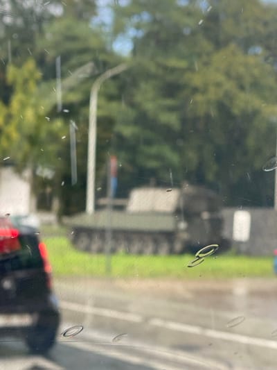 The image displays an outdoor scene viewed through a transparent surface, likely a window or glass pane, which is covered with numerous small water droplets and streaks, as well as some prominent circular water marks. The image appears to be rotated 90 degrees clockwise; if oriented vertically, it would show a landscape viewed from a static position.
In the foreground, the clear surface dominates, showing various imperfections like small white streaks, possibly dust or scratches, and distinct water marks. Some of these water marks are circular or ring-shaped, varying in size, with one prominent ring-shaped mark located towards the upper right of the image (or mid-right if rotated vertically). Subtle wavy patterns are also visible across the glass, suggesting reflections or textured glass.
Through this transparent surface, a blurry daylight scene is visible. On the left side (or top left if rotated vertically), there is a large area of dense green foliage belonging to trees, indicating a natural or landscaped environment. The trees are out of focus, creating a soft, hazy background. A patch of lighter blue, likely the sky, is visible through the top left portion of the trees.
Towards the right side of the image (or mid-ground if rotated vertically), a paved area, possibly a road or pathway, runs horizontally. Adjacent to this paved area, further to the right (or higher up), is a strip of bright green grass.
In the middle-to-far ground, a large, dark-colored vehicle or structure is partially visible. It has a robust, possibly industrial, appearance with visible vertical and horizontal lines, suggesting a truck, bus, or machinery. A lighter-colored rectangular panel is visible on its side. Further in the background, there are hints of blue and red colors, possibly from a sign or another distant object.
In the lower right corner of the image (or bottom right if rotated vertically), the rear section of another dark-colored vehicle, possibly a car, is visible. Its red taillights are illuminated.
The overall impression is that the photo was taken from inside a vehicle or building, looking out onto a road or street lined with trees and some structures, likely during the daytime. The numerous water marks suggest it might be raining, or has recently rained, or the window is simply wet or dirty. No discernible text is visible in the image.