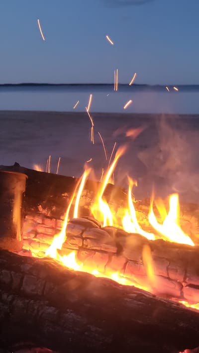 The image depicts a campfire burning on a beach at dusk. The fire is built on logs, with flames reaching upwards and sparks ascending into the twilight sky. In the background, a body of water is visible, with a thin horizon line separating it from the sky, which transitions from a deep blue to a lighter hue near the horizon. The beach appears sandy and damp, with some blurred figures in the distance. The overall atmosphere is serene, with the warm glow of the fire contrasting with the cool evening air. The location is identified as Zaostrov'e, Russia.
