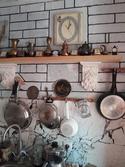 This is a medium shot, eye-level, indoor photograph of a kitchen wall with a shelf displaying various decorative items and kitchenware. The wall is covered in a white brick-patterned wallpaper with black grout lines. A wooden shelf runs horizontally across the upper part of the image, laden with an assortment of clocks and decorative objects, including vases and teapots. Above the shelf, a square clock with a cream-colored face and Roman numerals is mounted on the wall. To the left of the clock, a plastic bag hangs.

Below the shelf, the wall transitions to a marble-patterned wallpaper. Several kitchen items are suspended from hooks on this lower section of the wall. These include two frying pans, one larger and one smaller, a colander, and a small metal anchor. To the far left, a chrome faucet and sink are visible, with various cleaning supplies and containers. Further to the right, a small electrical socket with a plug is mounted on the wall.

The overall impression is of a well-used, somewhat cluttered kitchen, with a mix of antique and functional items. The lighting appears to be ambient, coming from an unseen source, casting subtle shadows. There are no people visible in the image. The style of the wallpaper and the collection of decorative items suggest a possible location in Cimișlia, Moldova, given the context.