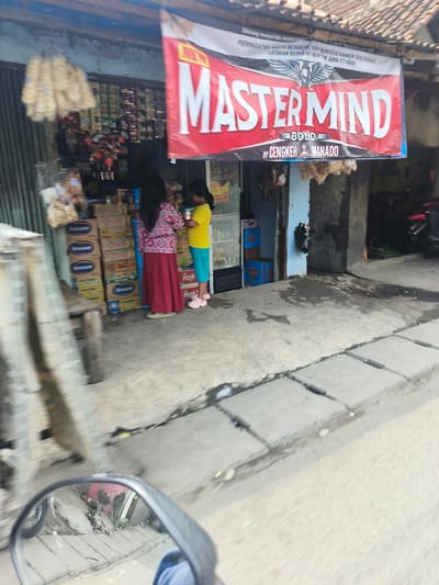 Two people stand outside a small shop with a "Master Mind" sign overhead. A woman in a floral top and red skirt and a girl in a yellow shirt and teal pants are near the entrance. The store has snacks and drinks displayed. Motorbikes are parked nearby. A street runs past the shop. A motorcycle mirror is visible.