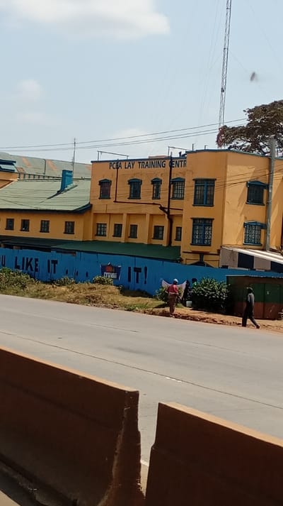The image displays an outdoor urban environment in Sigona ward, Kenya, under a clear sky with scattered clouds. In the foreground, two brown concrete barriers are partially visible. Beyond these barriers, an asphalt road with faint lane markings extends horizontally across the frame.
In the midground, a dirt verge with sparse dry vegetation separates the road from a blue barrier, possibly a corrugated fence or wall. This blue barrier exhibits white text, including "U LIKE IT" on the left and "IT!" on the right. Three individuals are present near this barrier. One individual, wearing a dark shirt and trousers, is walking right to left on the dirt verge. Another individual, dressed in a red shirt, dark trousers, and a light blue cap, stands facing the blue barrier, near what appears to be laundry on a line and a white bucket. A third person is partially visible behind the second individual, near a cluster of green bushes. To the far right, a small structure with dark green and brown sections is partially visible.
The background is dominated by a multi-story building painted yellow or ochre, featuring numerous windows with dark blue/green frames. The building's upper facade bears the inscription "PCBA LAY TRAINING CENTR." To the left of this main building, another structure with a dark green corrugated roof and yellow walls is visible. Power lines traverse the upper portion of the scene, and a tall, slender metal lattice tower stands prominently on the right side of the frame, extending towards the sky.