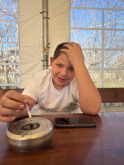 A child is indoors, positioned at a wooden table, holding a lit cigarette next to a metal ashtray containing cigarette butts. A smartphone is located next to the child. The background shows a window with a view of bare trees and a blue sky. Based on the window construction, it is likely the child is in some kind of tent or outdoor structure that is open or has large windows. The location may be Zlatna Panega, Bulgaria.