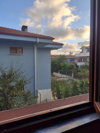This image captures a view from a window in Kandıra, Turkey. The scene depicts a residential area with several houses visible. In the foreground, a blue house with a reddish-brown tiled roof is prominent. A portion of a window frame, made of dark wood, borders the right side of the image, suggesting the photo was taken from indoors looking out.
Behind the blue house, other buildings with tiled roofs can be seen, along with trees and greenery. A garden area with what appears to be vegetable plants is visible in the middle ground. The sky is partly cloudy, with a mix of blue sky and fluffy white and yellowish clouds, illuminated by the soft light of what seems to be late afternoon or early evening. A chimney is present on the roof of the blue house, and a solar water heater is visible on the roof of another building in the background. There are no people or animals visible in the frame. The overall impression is a quiet, suburban residential scene.