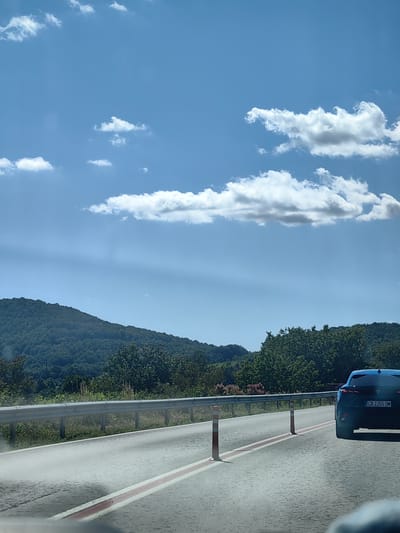 The image depicts a scenic view from a vehicle traveling on a road near Stefanovo, Bulgaria. The road is paved with visible red and white lane markings. A silver guardrail runs alongside the road, separating it from a grassy verge. Beyond the verge, a densely forested hillside rises, covered in lush green trees. In the distance, rolling hills are visible under a bright blue sky dotted with fluffy white clouds. A dark blue car with license plate "CB 2355 OM" is visible on the right side of the road, traveling in the same direction as the viewer. Orange and white traffic cones are placed at intervals on the side of the road. The overall impression is one of a clear, sunny day in a rural, mountainous region.