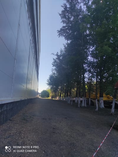 The image shows the side of a large, light-colored industrial building with a dark base. To the right of the building, a dirt path runs alongside a row of trees with whitewashed trunks. Beyond the trees, a large yellow piece of heavy machinery, possibly a bulldozer, is visible. The sky is clear and blue. The overall scene appears to be an outdoor industrial or construction area. The location provided, Kyzyl, Kazakhstan, does not appear to be visually verifiable from the image content itself, as there are no identifying landmarks or signs that can confirm this specific location.