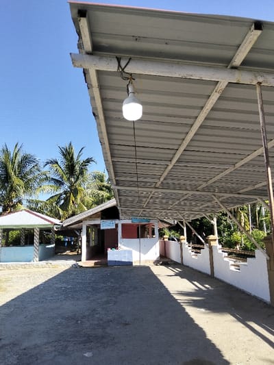 A low-angle view under a metal-roofed structure reveals a light bulb hanging from the ceiling by a cord. The structure's supports are visible. In the background are several buildings, painted in white, light blue, and red, with traditional designs. Palm trees are beyond the buildings. The ground is a mix of dirt and gravel with shadows cast from the roof. The sky is clear and blue.
