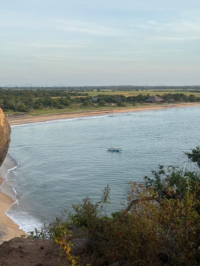 This image depicts a scenic view of a coastline in Shastrawela, Sri Lanka. The scene is dominated by the vast expanse of the ocean, its surface rippled by gentle waves. A small, traditional fishing boat with two occupants is visible in the water, suggesting a peaceful fishing activity or a leisurely boat ride. The shoreline is characterized by a sandy beach that transitions into lush greenery, with trees and vegetation lining the coast. In the background, hints of man-made structures, possibly resorts or dwellings with thatched roofs, are discernible amidst the foliage, indicating a developed or inhabited coastal area. The sky is overcast, suggesting a cloudy or misty day, likely early morning or late afternoon due to the diffused light. The overall atmosphere conveyed is tranquil and serene, highlighting the natural beauty of the coastal landscape. The image is taken from an elevated vantage point, possibly a cliff or a high ground, offering a panoramic perspective of the beach, ocean, and surrounding vegetation. The presence of a large rock in the foreground adds to the natural grandeur of the scene. There is no visible text in the image.