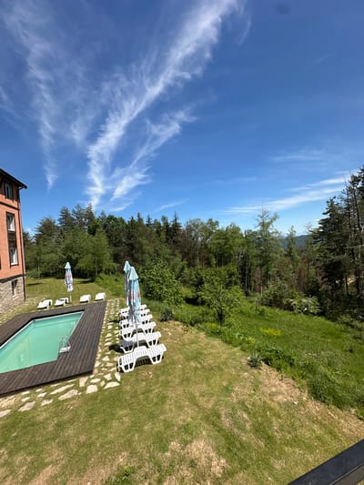 The image depicts an outdoor scene in Bogoslov, Bulgaria. A rectangular swimming pool with light green water is surrounded by a wooden deck and a stone pathway. White lounge chairs are arranged near the pool and under a blue and white striped umbrella. A lawn extends beyond the pool area, bordered by a dense forest of tall trees. The sky is blue with wispy white clouds. A building with orange walls and brown shutters is partially visible in the lower left corner of the frame.