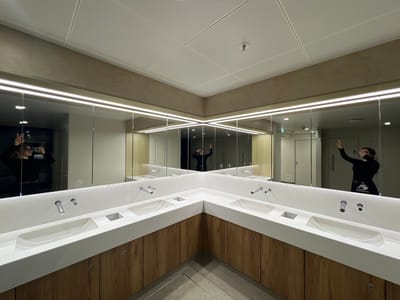 The image displays a modern, L-shaped public washroom located in Fischamend, Austria. A continuous strip of mirrors extends along two walls, reflecting the room and a single individual. Above the mirrors, a horizontal LED light strip illuminates the area. Below the mirrors, an L-shaped white countertop features four white, oval-shaped sinks, each with a wall-mounted chrome faucet. Small square recesses are present between the sinks. Underneath the countertop, wooden cabinets with horizontal grain are visible, fitted with small circular chrome handles. The ceiling is composed of white square panels, some exhibiting a ribbed texture, and a red fire sprinkler head is centrally visible. The floor is a light gray.
A female individual is reflected multiple times in the mirrors. She is wearing a black long-sleeved top, glasses, and dark lower garments with a white graphic detail on her right hip. She holds a black smartphone in her right hand, capturing her reflection, while simultaneously making a "peace" sign (V-sign) with her raised left hand. The reflections show plain white and dark doors in the background, suggesting other sections of the facility, and a black waste bin is partially visible in the far right reflection.