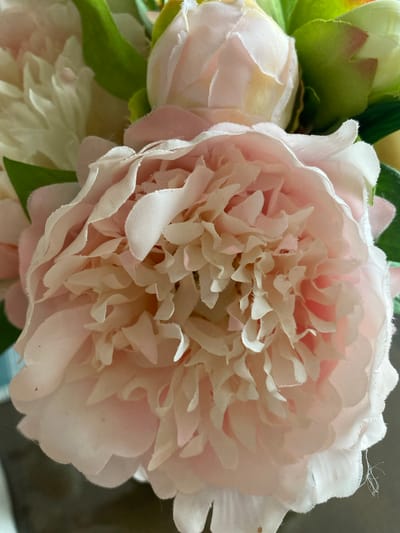 A close-up, eye-level shot shows a cluster of pink and white artificial flowers. The main subject is a large, pale pink peony in the foreground, with its many delicate petals unfurling. To the left and behind it are other flowers, including a white one and a partially visible pink bud. Green leaves are interspersed throughout the arrangement. The background is a blurry mix of neutral tones, suggesting an indoor setting. The lighting is soft and diffused, highlighting the texture of the fabric petals. This image does not contain any elements that would indicate its location in Les Cèdres, Canada, or any specific environmental context.