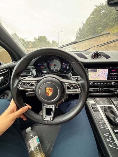 The scene is captured from inside a Porsche vehicle, on a rainy and overcast day. The driver's left hand grasps the black leather steering wheel at the 9 o'clock position, while a clear plastic water bottle rests on the driver's lap, covered by dark blue pants. The vehicle's speedometer indicates 61 mph. The central infotainment screen displays a navigation map with "Newton Street" clearly labeled, situating the location within Newton, United States. The screen also shows "107.9 FM HD1." Rain is visible on the windshield, with active wipers. Ahead on the multi-lane roadway, several vehicles are observed in the distance. The right side of the road is bordered by a concrete barrier, behind which a wooded embankment rises. Overhead streetlights are present along the road.