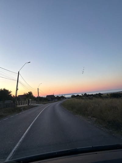 The image depicts a road in a rural area, possibly near Pașcani, Romania, during sunrise. The sky transitions from a pale blue at the top to hues of orange and pink near the horizon, indicating the early morning. A paved road curves through the scene, with a white line marking its center. On the left side of the road, there are houses with roofs and fences, along with utility poles and a complex network of wires. Tall streetlights are also visible along the roadside. The right side of the road is bordered by dry, tall grass and scattered trees. In the distance, a thin layer of fog or mist hovers over a body of water, possibly a river or lake. The image appears to be taken from inside a vehicle, as the bottom of the windshield is visible. There are no people or animals in the frame. The overall atmosphere is peaceful and serene, characteristic of a rural dawn.