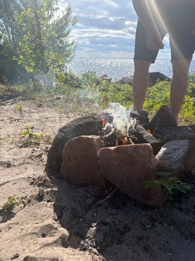 The image depicts a campfire on a sandy beach near a body of water, likely the Kyiv Reservoir, in Kozarovychi, Ukraine. A person is partially visible in the background, standing with their legs exposed from the knees down, wearing dark shorts. The campfire is in the foreground, surrounded by rocks, with burning wood and wisps of smoke rising into the air. The sun is shining brightly, casting lens flare effects across the image, and the sky is partly cloudy. Vegetation and trees are visible on the left side of the frame, bordering the beach. The ground is covered with sand, scattered twigs, and some small green plants.