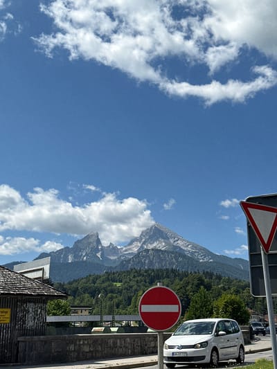 The image depicts a sunny day in Berchtesgaden, Germany, with the Watzmann mountain range dominating the background. In the foreground, a white Volkswagen Touran is parked on the right side of a road. To the left of the car, a red circular sign with a white horizontal bar indicates "no entry." A triangular "yield" sign is visible on the right side of the road. A small wooden building with a dark, shingled roof is situated on the left side of the image. The sky is a bright blue, with scattered white clouds. The overall scene suggests a picturesque mountain town setting.