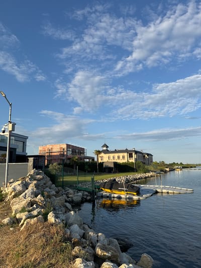 This is a daytime, eye-level shot of a waterfront in Kazashko, Bulgaria. The scene is dominated by a body of water on the right, reflecting the bright blue sky with scattered white clouds. On the left, a rocky embankment lines the shore, leading up to a modern house with a light grey facade and large windows. Next to it, a construction site is visible with a building covered in scaffolding. Further down the shore, a large, ornate building with a dark roof and beige walls stands prominently. A yellow personal watercraft is docked at a floating pontoon. In the foreground, dry grass and large rocks are scattered along the edge of the embankment. A lamppost with attached security cameras stands on the left side of the frame. The overall impression is of a serene, semi-developed residential area.