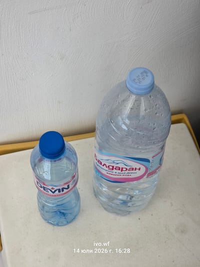 Two plastic bottles of water are placed on a light-colored, textured surface. The bottle on the left is smaller and has a blue cap with the brand "DEVIN" visible on its label. The bottle on the right is significantly larger, also with a blue cap. This bottle has condensation on its surface. Its label features the word "Балдаран" in red and a pink and blue design. A yellow ledge is visible in the background, behind the bottles. The overall environment appears to be indoors, with a plain, textured white wall behind the yellow ledge. The bottom of the image contains text that reads "ivo.wf 14 юли 2026 г. 16:28", indicating a date and time. No people are present in the image. The image does not provide any information about the city of Chal, Bulgaria.