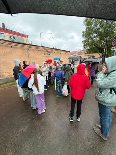 A group of people, mainly children, stand in a line on a wet asphalt road in what appears to be Tavry, Russia. Some hold umbrellas, and most are wearing coats or jackets. The line stretches along the side of a building constructed from orange bricks. A sign in Russian with a portrait is visible on the side of the building. The sky is overcast.