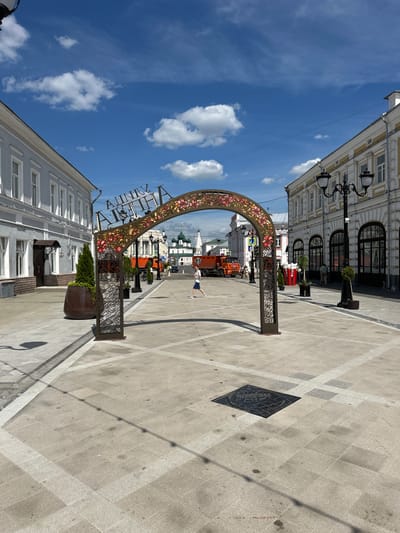 The image depicts a street scene in Putyatino (Semivragovskaya S/A), Russia, on a sunny day. The sky is bright blue with scattered white clouds, indicating clear weather. The perspective is a low-angle shot, looking upwards towards buildings and the sky.
The primary subject is an ornate archway adorned with floral patterns, possibly an entrance to a pedestrian zone or a decorative landmark. The arch is metallic and appears to be a prominent fixture in the urban landscape. In the background, traditional Russian architecture with multiple windows and classical detailing lines the street. Lampposts with multiple globes are also visible, suggesting a historic or well-maintained urban area.
A person is seen in the distance, appearing to walk or pose near the arch. Further down the street, a construction or maintenance vehicle, possibly an orange truck, is parked. Planters with greenery are placed along the sidewalk, adding to the street's aesthetic. The ground is paved with large, light-colored tiles arranged in a patterned grid, with some darker square inlays and a diamond-shaped metal grate visible.
The time of day appears to be midday, given the strong sunlight casting distinct shadows. The overall mood is bright and cheerful, characteristic of a pleasant day outdoors in a city. The visible text on the arch appears to be in Cyrillic script, but it is partially obscured and difficult to read clearly. The scene captures a moment of urban life, with elements of architecture, public art, and everyday activity.
