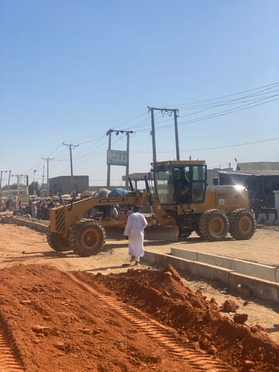 The image depicts an active road construction scene in Rijiyar Lemo, Nigeria, under a clear blue sky. In the foreground, reddish-brown earth with visible tire tracks covers the ground. A large, yellow SANY SY3165C motor grader, with six substantial off-road tires, occupies the mid-ground. Its blade is positioned low, and an operator wearing a yellow high-visibility vest is visible within the cabin. Directly in front of the grader's rear section, an individual dressed in a white, full-length garment and light-colored footwear stands with their back to the viewer, facing the grader.
To the right of the grader, recently installed concrete drainage channels or curbstones are visible, running parallel to the unpaved road. In the background, numerous utility poles with multiple power lines extend across the scene. A prominent sign displaying "DTH" and an eyeglasses graphic is mounted on a pole to the left of the grader. Various single-story and multi-story structures, some with corrugated metal roofs, line both sides of the road. On the far left, a cluster of people and what appears to be an informal market or street vendor setup is discernible. The scene is brightly lit by natural sunlight, with a glare visible on the grader's metallic surfaces.