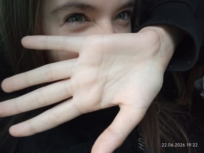 A close-up, selfie-style shot shows a person's face and hand. The person's eyes, with visible eyelashes and eyebrows, are looking towards the left side of the frame. A left hand is held up in front of the lower half of the face, obscuring the nose and mouth. The palm of the hand is visible, with fingers extended. The person appears to be wearing a black garment, possibly a hoodie, with a visible sleeve on the right side. Long, dark brown hair is visible framing the face. The background is dark and out of focus. A timestamp in the bottom right corner reads "22.06.2026 19:22". The location of Sarytogay, Kazakhstan is not depicted in this image.