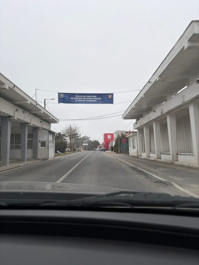 The image depicts a straight-on view from the interior of a vehicle, looking forward towards the Vama Veche border crossing in Romania under an overcast sky. A two-lane asphalt road with a solid white center line extends into the distance. Two substantial, multi-columned, flat-roofed concrete structures, typical of a border control station, flank the road on both the left (north) and right (south) sides.
Spanning overhead between these structures is a dark blue banner. The banner features the emblem of the Romanian Border Police on the left and the flag of Romania on the right, with white text in Romanian reading: "POLITIA DE FRONTIERA VĂ DOREȘTE CĂLĂTORII PLĂCUTE ÎN SPATIUL SCHENGEN!" (Border Police Wishes You Pleasant Journeys in the Schengen Area!).
On the left, beyond the main building, a smaller white booth-like structure is visible, followed by a gray gate or barrier in the distance. Sparse, mostly bare trees line the left side of the road. On the right, past the main building, a red circular 'no parking/stopping' sign is visible, along with orange traffic cones lining the shoulder. Further down on the right, a prominent red building with white accents is present. Several vehicles are parked or stationary further down the road in both directions. The road surface exhibits some wear and minor undulations, particularly on the right shoulder. No individuals are clearly discernible in the immediate foreground.