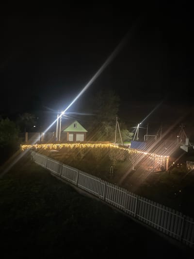 The image depicts an outdoor scene at night in what appears to be the city of Хлябово, Russia. A small house with a green roof is visible, illuminated by a bright light source that causes significant lens flare. A fence adorned with string lights runs alongside the house. In the background, there is a structure with a blue roof supported by wooden posts, also lit by string lights. A slatted bridge or walkway is seen on the right side of the frame. The surroundings appear dark, with only minimal ambient light. The image is rotated counter-clockwise.