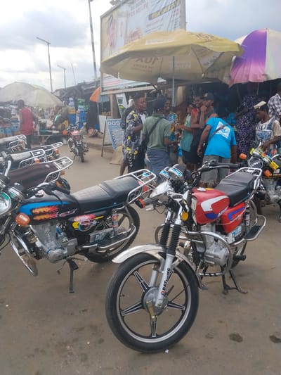 The image depicts a bustling street scene in Umu Enge, Nigeria, with several motorcycles parked on the pavement. A group of people are gathered under a yellow umbrella near a makeshift market stall. Signage includes advertisements for "POS Lalu Provision Store" and a "BURIAL ARRANGEMENT." The weather appears overcast.