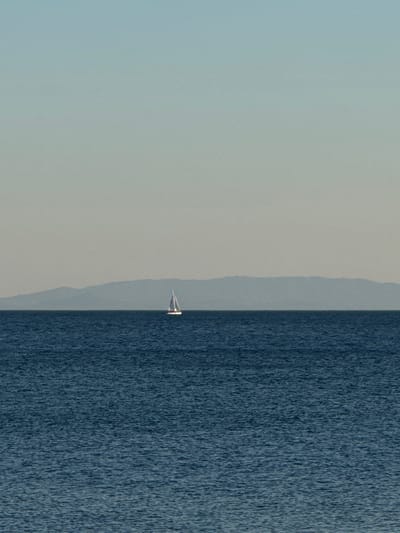 A sailboat with a white sail is pictured in the distance on the sea, with a hazy mountainous landmass in the background. The sea is a deep blue, with subtle ripples on its surface. The sky above is a pale, gradient blue, transitioning to a lighter hue near the horizon. The scene is serene and expansive. The location is the coast of Alimos, Greece.
