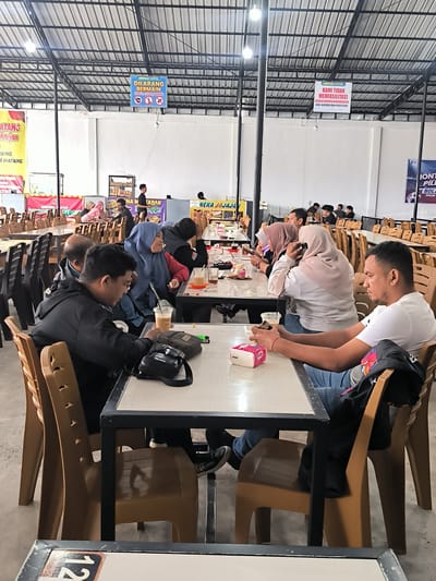 The image depicts an indoor eating area with multiple tables and chairs, likely in Panteraya, Indonesia. Several groups of people are seated at tables, engaged in eating, drinking, and conversation.

In the foreground, a man in a white t-shirt and jeans is seated at a table, looking towards the right. He is holding a mobile phone in his left hand and has a black jacket draped over the back of his chair. Next to him, a man wearing a black jacket and jeans is looking down at his phone. In front of him is a plastic cup with a drink.

Further back, a group of four individuals is seated at a table. Two women are wearing hijabs, one light pink and one blue. The woman in the blue hijab is looking to her right and holding a phone. The man seated next to her is wearing a black jacket. Another man is partially visible behind them, wearing a black jacket.

In the background, more tables and chairs are set up, and several other individuals are seated. Signs are visible on the wall and ceiling, including one that reads "DILARANG BERMAIN KARTU / DOMINO" (Forbidden to Play Cards/Dominoes) and another stating "KAMI TIDAK MEMFASILITASI SEGALA BENTUK KEEMAKSIAN (JUDI, NARKOBA DAN PROSTITUSI)" (We do not facilitate any form of vice (gambling, narcotics, and prostitution)).

The environment appears to be a covered outdoor or semi-open space, with a metal roof structure and white walls. The lighting suggests it is daytime. The overall scene is that of a casual dining or gathering space.