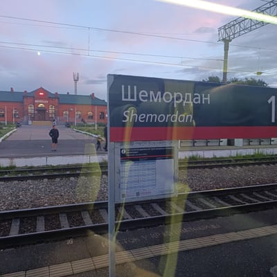 This image depicts a train station in Shemordan, Russia, during twilight. The main subjects are the train station building, railway tracks, and a signpost displaying train schedules.
The station building is constructed of red brick with green roofing, featuring arched windows and a prominent sign above the entrance that reads "ШЕМОЛДАН" (Shemordan) in Cyrillic. The signpost in the foreground also displays "Шемордан" in both Cyrillic and Latin alphabets, alongside "Timetable suburban trains," indicating it's a functional railway stop. A train timetable is visible on the signpost, listing departure and arrival times and destinations.
In the scene, a few people are visible. One man in a black jacket and shorts stands on the platform, looking towards the station. Another person, possibly a child, is walking in the background. The railway tracks run across the foreground, with ballast and concrete sleepers clearly visible. The sky is painted with the soft hues of dusk, with clouds and a faint moon. The overall atmosphere is calm and peaceful, suggesting the end of a day. Reflections on what appears to be a window can be seen, overlaying parts of the scene and the sign. The image captures a typical moment at a provincial Russian train station.