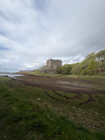 The image shows Eilean Donan Castle, a 13th-century castle situated on a small tidal island in Loch Duich, in the Scottish Highlands. The castle is visible in the center of the frame, a formidable stone structure with turrets and a flag flying atop. The surrounding landscape is characterized by a dramatic coastline, with rocky shores, green hills, and calm water of the loch. The sky is overcast, with white and grey clouds dominating the scene. The perspective of the image suggests a ground-level view, possibly taken from a nearby shoreline.