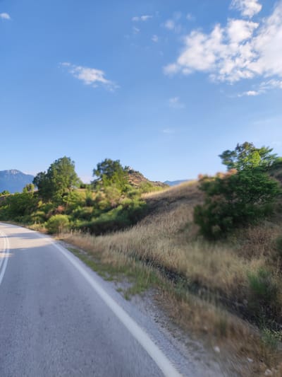 The media file displays a scenic view of a road winding through a hilly, verdant landscape under a clear blue sky with scattered white clouds. The time of day appears to be late afternoon or early evening, indicated by the soft sunlight casting shadows and highlighting the textures of the vegetation. The weather is pleasant, with a bright and sunny disposition.
The scene features a paved road with white lane markings, suggesting it's a route used for travel. The road occupies the left side of the frame, appearing to curve away from the viewer. Adjacent to the road, the terrain rises into a hillside covered in dry grasses, shrubs, and scattered trees. These trees, varying in size and density, contribute to the lush, natural feel of the environment. In the distance, a mountain range is visible, adding depth and grandeur to the landscape. A small structure, possibly a building or monument, is perched on the crest of the hill, a subtle focal point amidst the natural scenery.
The overall impression is one of tranquility and natural beauty, characteristic of a rural or mountainous region. The slightly blurred foreground and midground, possibly due to motion, suggest the image was taken from a moving vehicle. There is no visible text in the media file. The location context, Μεγάλη Κερασιά, Greece, points to a specific geographical area known for its natural landscapes.
