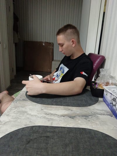 A young man with a buzz cut is seated at a table, looking down at his phone. He is wearing a black t-shirt with a graphic design on the front. He holds a white mug in his left hand, with a spoon inside. The table has a marble-like pattern and is covered with a dark placemat. To his right, on the table, is a black container filled with individually wrapped candies and some pastries in a plastic bag. A white box with blue and black graphics is also visible on the right side of the table. The background shows a wall with vertical stripes, a closed closet door on the left, and a large cardboard box leaning against the wall. A chair with a purple cushion is behind the man. The lighting appears to be artificial, suggesting an indoor setting, possibly a home. There is no indication of the time of day or weather conditions. The man's facial expression is neutral, focused on his phone. The scene takes place in Ильинка, Russia.