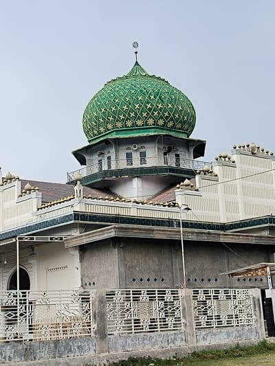 This image captures a detailed exterior view of a mosque in Grong-Grong, Indonesia, under an overcast sky. The central and most striking feature is a large, vibrant green dome, adorned with intricate gold-colored geometric patterns that include stylized stars and crisscross designs. A slender, golden finial extends from the dome's peak, crowned with the symbolic crescent moon and star.

Below the main dome, the mosque features a multi-level structure. A circular section directly beneath the dome incorporates small balconies with ornate white railings and windows, indicating an upper floor or a balcony area. Further down, terracotta-tiled roofs are visible, interspersed with white wall sections that are topped with numerous small, decorative, bulb-shaped finials, some appearing golden. These sections also feature vertical ridging and dark green and gold decorative friezes.

The lower visible parts of the mosque combine white plastered walls with patterned textures and plainer concrete sections. In the foreground, a decorative white metal fence with intricate diamond and geometric patterns runs horizontally across the frame, partially obscuring the base of the building. Behind the fence, a gray concrete wall is visible, with a strip of sparse grass and dirt at the very bottom of the frame. A single street light pole can be seen on the right side, extending from behind the fence upwards.

The image is captured during the daytime, characterized by soft, diffused lighting consistent with an overcast or cloudy sky, casting no harsh shadows. No people or specific activities are visible, presenting a static architectural portrait of the mosque.