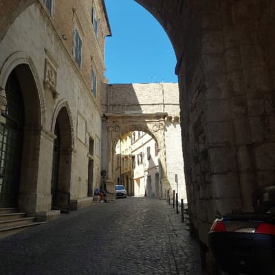 The image depicts a historical street scene in Ancona, Italy, specifically Via della Loggia looking towards the Arco dell'Ammiragliato (also known as Arco Bonarelli). The foreground is dominated by a large, dark stone archway on the right, which casts a deep shadow across the immediate street. The rear portion of a dark vehicle, possibly a scooter or small car, with prominent red taillights, is partially visible in this shadow. Several dark, vertical bollards line the right edge of the cobblestone street.

On the left, a multi-story historical building constructed of light-colored stone with a brick upper section features multiple arched entrances at ground level, some containing dark green doors. Two individuals are seated on a stone ledge at the base of this building. One person wears a dark top and light-colored lower garments, while the other is partially obscured.

Further down the cobbled street, a white compact car is parked. Beyond the car, the ornate stone Arco dell'Ammiragliato spans the street, acting as a gateway. Buildings beyond this archway, also constructed of stone and featuring windows with shutters, are visible under direct sunlight. The sky above is clear and bright blue. Strong sunlight illuminates the background and parts of the midground, contrasting with the deep shadows in the foreground.