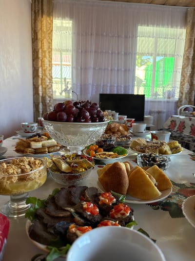An indoor scene in a residence in Урмаево, Russia, displays a large dining table prepared for a meal under natural daylight. The table is covered with a white tablecloth featuring a red and green floral pattern. A prominent crystal bowl in the center-left contains a substantial quantity of red grapes and cherries. Surrounding this centerpiece are numerous plates and bowls filled with a variety of food: golden-brown pastries and cakes, small wrapped candies with Cyrillic text visible on some labels, assorted dried fruits (orange, green, and dark), plates of sliced dark meat garnished with lettuce and small appetizers topped with diced red vegetables and a white sauce, and triangular golden baked goods. Several teacups with saucers, some featuring red polka dots, are arranged on the table. In the background, two windows are visible, adorned with sheer white lace curtains layered under heavier gold-patterned drapes. Through the windows, a blurred exterior view shows greenery, parts of building roofs, and a distinct bright green structure. A dark, rectangular object, possibly a turned-off screen, is positioned between the windows, and a white heating radiator is partially visible beneath the right window. The room features light-colored walls and wooden trim along the ceiling. No individuals are present in the image.