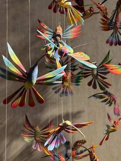 A collection of brightly colored, intricately carved wooden hummingbird figures are suspended from thin horizontal strings against a neutral-toned background. The hummingbirds display a variety of vibrant hues, including blues, reds, greens, and yellows, arranged in feather-like patterns. Their wings are spread, creating a dynamic sense of flight and movement within the static display. The lighting suggests an indoor setting, possibly a museum or gallery.