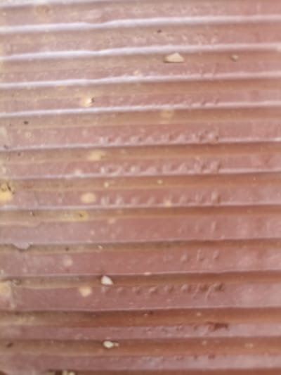 The image shows a close-up, out-of-focus view of a reddish-brown, corrugated surface. The surface has horizontal ridges and appears to be weathered, with some lighter-colored specks and darker marks scattered across it. The overall texture is uneven. There are no people or discernible objects within the frame that would suggest a specific location in Mahmudlu, Azerbaijan, or any other specific context. The image solely focuses on the abstract texture of the material.