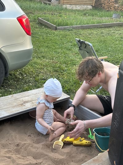 The image depicts a young child in a blue and white striped outfit and white bonnet sitting in a sandbox. An older person, shirtless with curly brown hair and glasses, is kneeling next to the sandbox, engaging with the child. In the sandbox are several yellow plastic toys including a shoe, a bucket, and a shovel. A car is visible on the edge of the sandbox. The setting is outdoors, in a grassy area. The location is Пальники, Russia.