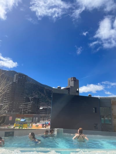 The image shows a view in Les Escaldes, Andorra, from a building with a swimming pool. A clear blue sky with scattered white clouds fills the upper left portion of the frame. A mountain covered with dense trees rises vertically in the center. Modern buildings stretch along the base of the mountain, blending into the urban environment. The right side of the image features a swimming pool with several people in the water. A building is adjacent to the pool.