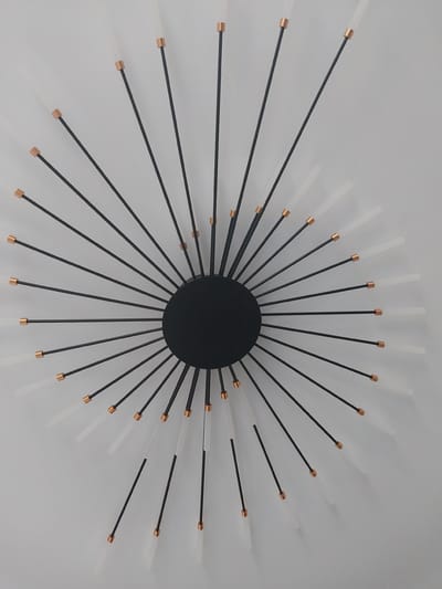 The image shows a modern sunburst-style wall clock with a black circular center. Thin black rods radiate outwards from the center, tipped with copper accents and clear or frosted glass elements. The clock is mounted on a plain white wall. The image appears to be taken from directly above. This object is located in Nizhneye Myachkovo, Russia.