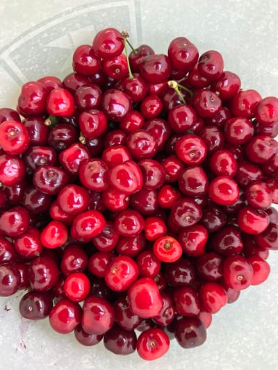 The image shows a close-up of a pile of fresh, red cherries. The cherries are densely packed together, filling most of the frame. Their surfaces are glossy, reflecting light, and their colors range from bright red to deep burgundy. A few cherries still have stems attached. The background is a light gray surface. The location in the city of Goran, Bulgaria is not evident.