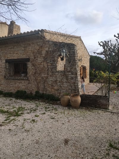 The image depicts the exterior of a stone house in Aubagne, France, under an overcast sky. The foreground consists of light-colored gravel with sparse patches of green grass.

The single-story building features walls constructed from irregularly shaped, light-colored stone blocks. The roof is tiled, with a dark gutter system running along its edge. A stone chimney structure is visible on the left side of the roofline. A bare vine with tendrils covers portions of the stone wall.

A rectangular window with a dark, decorative wrought-iron grill is prominent on the left side of the house's visible facade. To its right, an arched recess in the stone wall features a dark, ornate lantern affixed to a wooden support. Further to the right, a second, smaller dark lantern is mounted on the wall. A partially open wooden shutter is visible on an opening to the far right, revealing an interior or a darker recess.

In front of the house, near the wall, two large, weathered earthenware pots are placed on the gravel. A low strip of green vegetation runs along the base of the wall.

On a paved patio area extending from the right side of the house, a single individual, appearing to be a young person, is visible from behind. They are wearing light blue trousers and a light-colored long-sleeved top, standing or moving towards the right. A dark, woven barrier or planter box separates the gravel from the patio on the lower right.

In the background, a large, leafless, gnarled tree is visible on the right, with dense green trees further behind it. Bare tree branches are also visible against the sky on the far left of the frame.