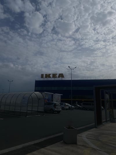The image shows the IKEA store in Bielany Wrocławskie, Poland. The store is a large, blue building with the IKEA logo in large orange letters on the facade. In front of the store is a parking lot with several cars and a covered walkway for shopping carts. The sky is cloudy, with a mix of white and gray clouds. The lighting suggests it is daytime. The scene is presented in a static, observational manner.