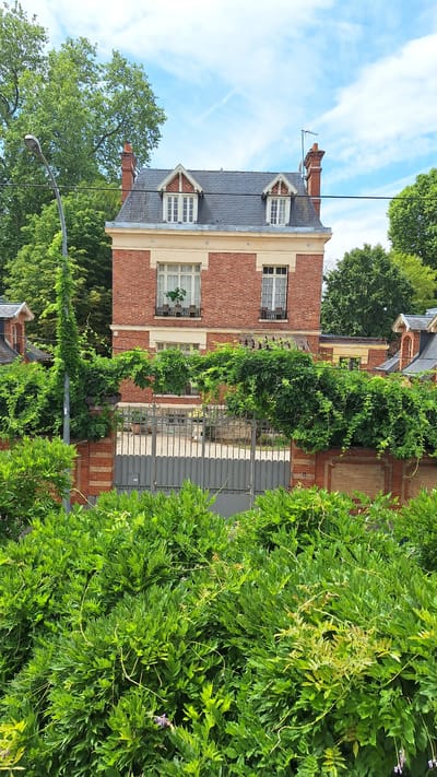 The image depicts a two-story brick house with a gray slate roof, located in Fontainebleau, France. The house features dormer windows with white trim and an ornate brick chimney. A modern gray metal gate stands in front of the house, flanked by brick walls and overgrown green foliage. A streetlamp covered in ivy is visible to the left of the house, and lush trees surround the property. The sky is partly cloudy with blue hues. The overall scene suggests a well-maintained residential property in a verdant setting.