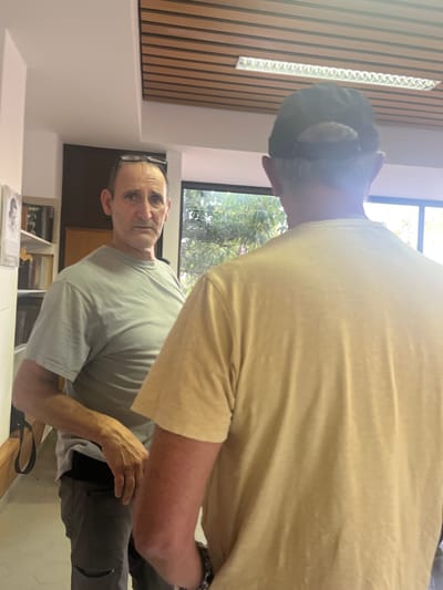 The image shows two adult males standing indoors in a location identified as the Brener Regional Council, Israel. The setting features a ceiling with wooden slats and recessed linear fluorescent lighting. To the left, a white bookshelf holds books and a framed picture. A large window on the right provides a view of green foliage outside.
In the foreground, a male subject is seen from behind, wearing a light-colored, short-sleeved t-shirt and a dark baseball cap worn backwards, revealing gray hair at the nape of his neck. A metallic chain-link bracelet is visible on his right wrist.
Behind him and slightly to the left, a second male subject faces the camera, gazing directly forward with a neutral expression. He wears a light grey, short-sleeved t-shirt and dark-rimmed eyeglasses perched on his forehead. His lower body shows dark grey pants with a distressed area on the left knee, and a black strap hangs near his left hip. No direct physical interaction between the two subjects is depicted.