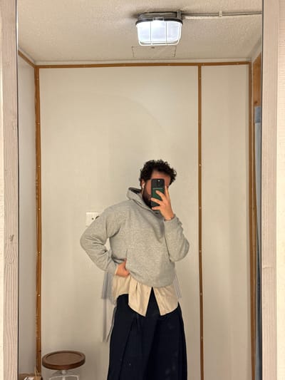 This image depicts a male individual's reflection in a mirror, located in a fitting room or similar private space within the city of Gondomar, Portugal.
**Subject:** One adult male, seen from the mid-chest to mid-shin. He possesses dark, curly hair and a beard, with his face obscured by a smartphone held to take the photograph. He wears a white AirPod in his visible left ear and a silver-colored bracelet on his left wrist.
**Attire:** The individual is dressed in a light grey hooded sweatshirt, worn over a light beige collared shirt. A light-colored, possibly striped, garment is visible beneath the beige shirt on the left side. He wears wide-leg, dark blue or black trousers with faint vertical pinstripes.
**Action/Interaction:** The male subject is standing with his left hand on his hip, while his right hand holds a dark green smartphone, featuring a dark wallet or card holder attached to its rear, directly in front of his face. This indicates a self-portrait (selfie) being taken.
**Environment:** The setting is an enclosed room, likely a fitting room, characterized by plain, light-colored (off-white or light beige) walls. These walls are segmented by thin, light brown wooden strips with visible small, round fasteners, forming paneling. A rectangular mirror occupies the frame, with its reflection revealing a light-colored, textured ceiling featuring a single industrial-style cage light fixture and visible electrical conduit. A small, round wooden stool with a white metal base is positioned in the lower-left foreground of the reflection. A rectangular light switch or electrical outlet cover is present on the wall behind the subject's left arm.