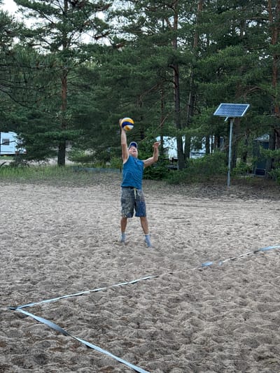 A man in a blue tank top and patterned shorts is playing volleyball on a sandy court. He is in mid-air, reaching up to hit the ball, which is above his head. He is wearing a blue baseball cap and grey socks. The volleyball court is marked with white tape on the sand. In the background, there are pine trees, a solar panel on a pole, and a recreational vehicle. The location is the city of Ozerki (Krasnodolinskaya), Russia.